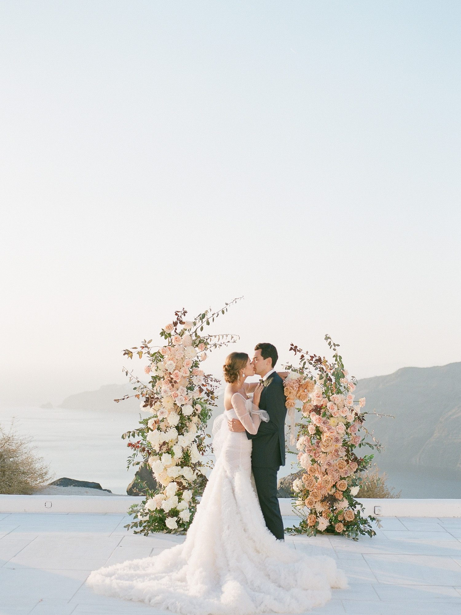 Bride and groom share a romantic kiss at sunset in Santorini during golden hour, overlooking the Aegean Sea.